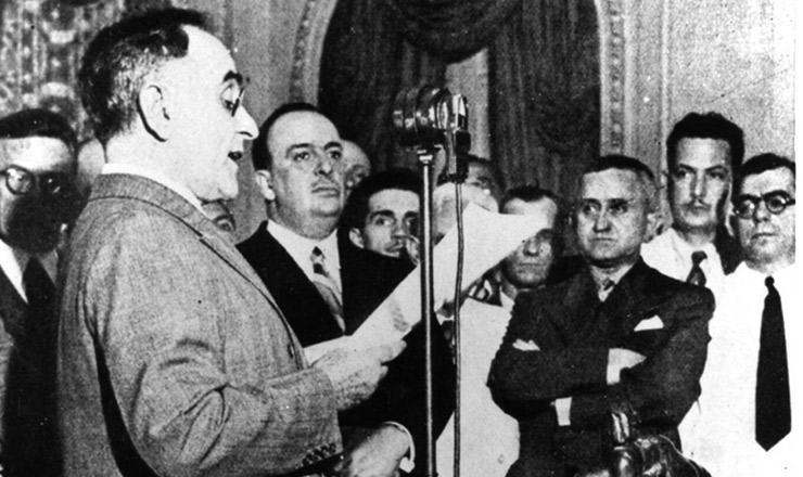  <strong> Getúlio Vargas anuncia </strong> pelo rádio o início novo regime ditatorial. À sua direita aparecem o ministro Dutra (de braços cruzados), Filinto Müller (de bigode, atrás dele) e o autor da Constituição, Francisco Campos (extrema direita)